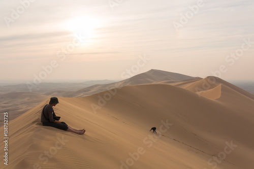 One person sits on top of the dunes of the desert, and the other climbs to the top