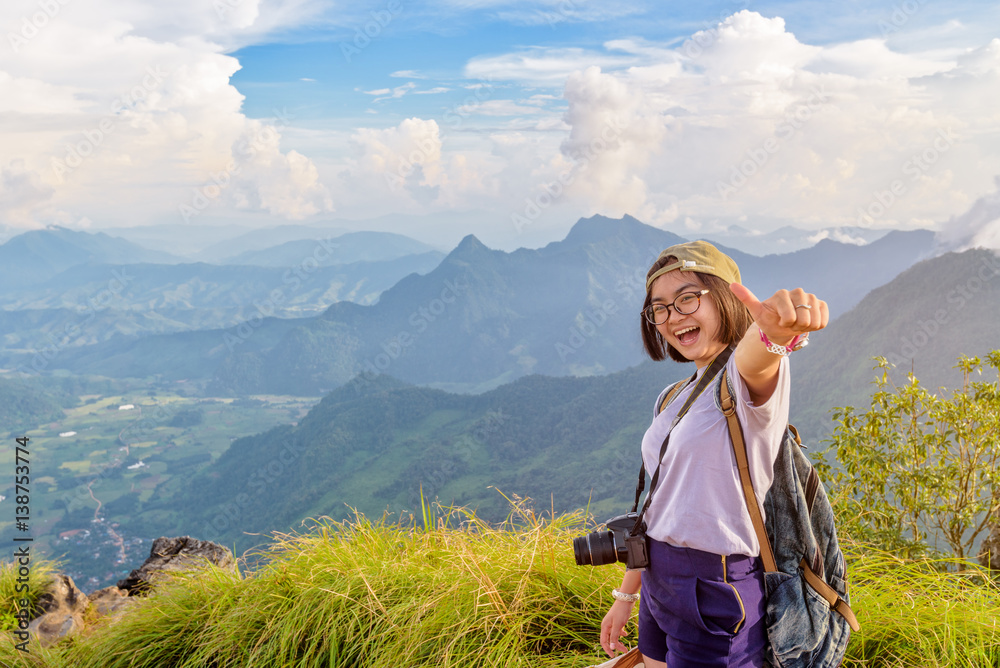 Naklejka premium Happy hiker asian cute teen girl with dslr camera cap eyeglass and backpack stand smiling poses thumb up on top of mountain at viewpoint Phu Chi Fa Forest Park in winter, Chiang Rai, Thailand