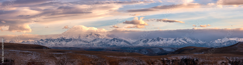 Panoramic view of a vivid sunset over Great Caucasian Ridge. Warm autumn light glows clouds and ice of snow caps of high Elbrus mountain peak and tops of other mountains. Gumbashi pass