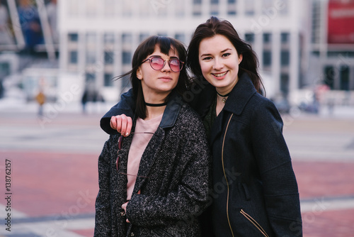 Outdoor portrait of two young, happy, beautiful and smiling girls in sunglasses. Girls in a dark dress on a background of the urban landscape.