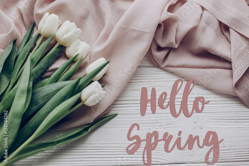 hello spring text fresh sign. stylish white tulips on beige soft fabric and rustic wooden table background top view. flat lay. soft light, tenderness moment.