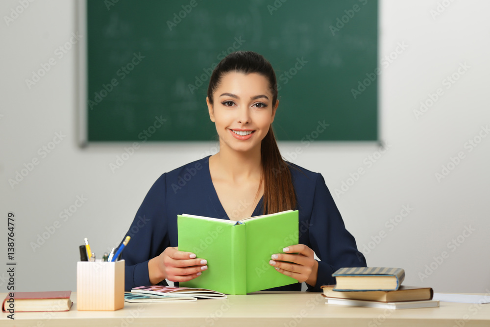Beautiful young teacher sitting at table in classroom Beautiful young teacher sitting at table in classroom