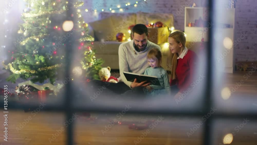 Looking Through the Window. Sitting Under Christmas Three with Gifts Under It Father and Mother Read a Book to Little Daughter. Shot on RED Cinema Camera 4K (UHD).