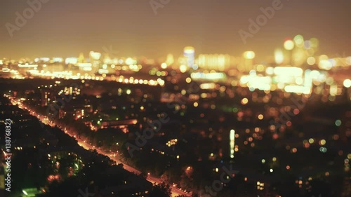 Time lapse tilt shift shooting from high above of night street in residential city district with trees, houses, working lanterns and moving cars, strong bokeh on horizon