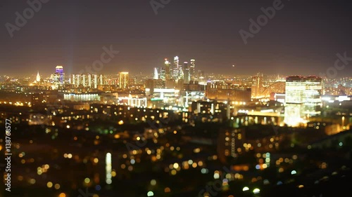 Time lapse tilt shift shooting from high point of night in big modern city with many lights and strong bokeh in foreground, clear sky with multiple flying airplanes, business skyscrapers in distance