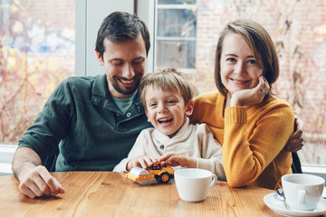 Portrait of white Caucasian happy family of three mother, father and son, sitting in restaurant cafe at table, smiling playing, authentic lifestyle