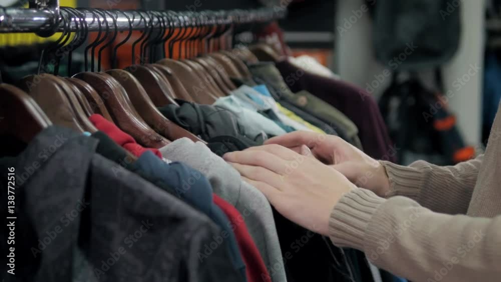 Men sweaters and shirts in different colors on hangers in a retail clothes store, man choose hoodies close up in shop Men sweaters and shirts in different colors on hangers in a retail clothes store, man choose hoodies close up in shop