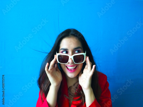 Shot portrait of surprised smiling woman whit long brunette hair and in white sunglasses in vibrant blue background with red clothes