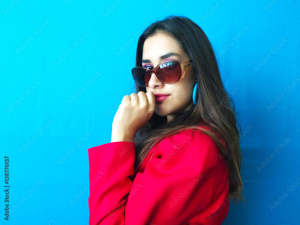Fototapeta premium Fashion sexy model in sunglasses, beautiful young woman with hand in her mouth with red coat clothes and long brunette hair in a bright vibrant blue color