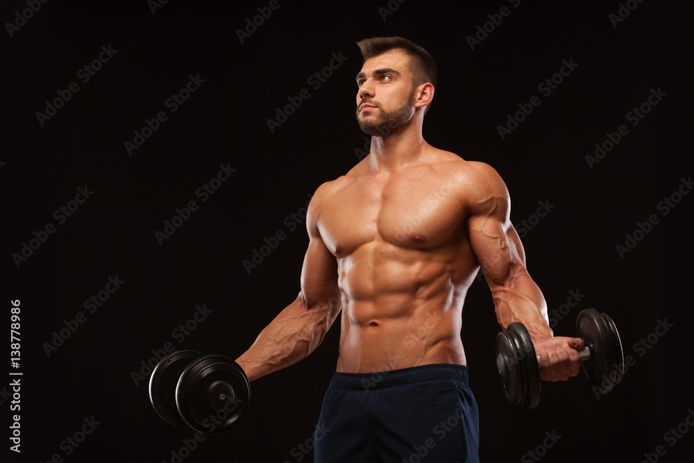 Handsome athletic man in gym is pumping up muscles with dumbbells in a gym. Fitness muscular body isolated on dark background.