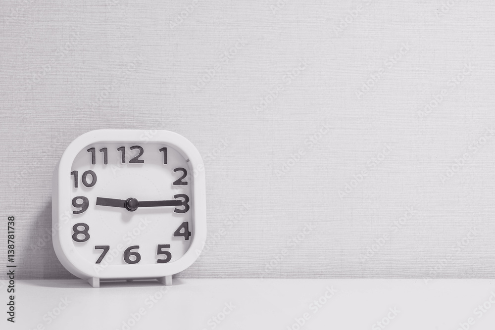 Closeup white clock for decorate show a quarter past nine or 9:15 a.m. on white wood desk and cream wallpaper textured background in black and white tone with copy space