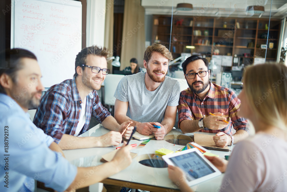 Fototapeta premium Group of young men sitting close together at table during meeting in modern spacious office, listening intently to their female boss, suggesting ideas and discussing data