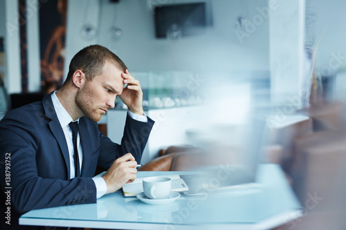 Young handsome man wearing formal attire working with laptop in empty hall of modern restaurant during coffee break, looking puzzled and thinking hard trying to solve some important matter