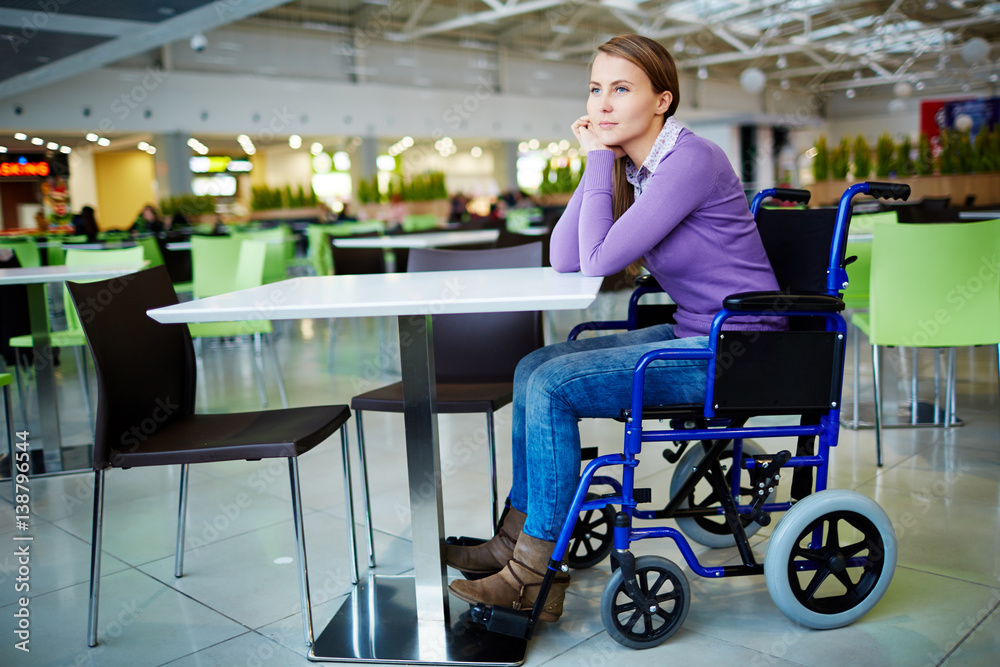 Young physically impaired girl sitting in wheelchair at mall food court and waiting for her friend with pensive face expression