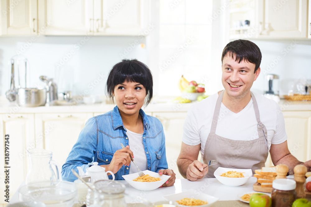 Attractive mixed race woman and her Caucasian husband in apron having lunch together in lovely kitchen