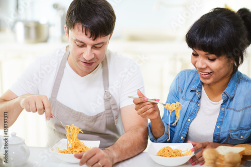Waist-up portrait of lovely multi-ethnic couple sitting at dinner table and eating spaghetti with appetite