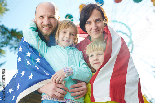 Portrait of patriotic family with American flag looking at camera with wide smiles while celebrating Independence Day in park