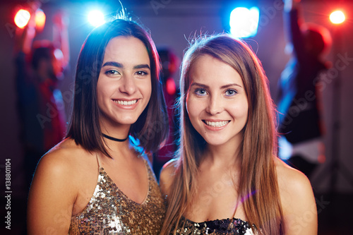 Portrait of two young pretty girls in shining tops looking at camera with bright toothy smiles while hanging out in nightclub
