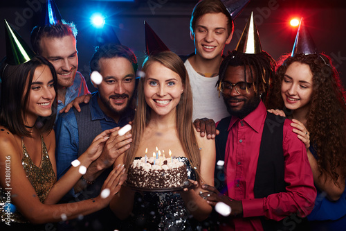 Group photo of dressed-up young people celebrating birthday of their female friend standing in the middle with cake and smiling happily