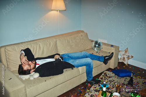 Canvas Print Asian man in jeans and jacket having nap in messy living room after wild New Yea