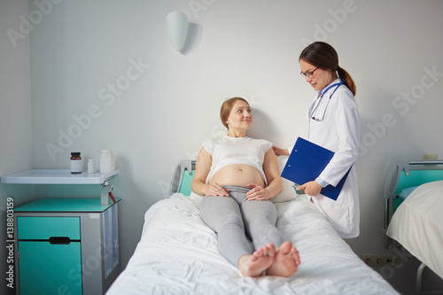 Young pregnant woman lying in bed in hospital room and listening to her doctor attentively