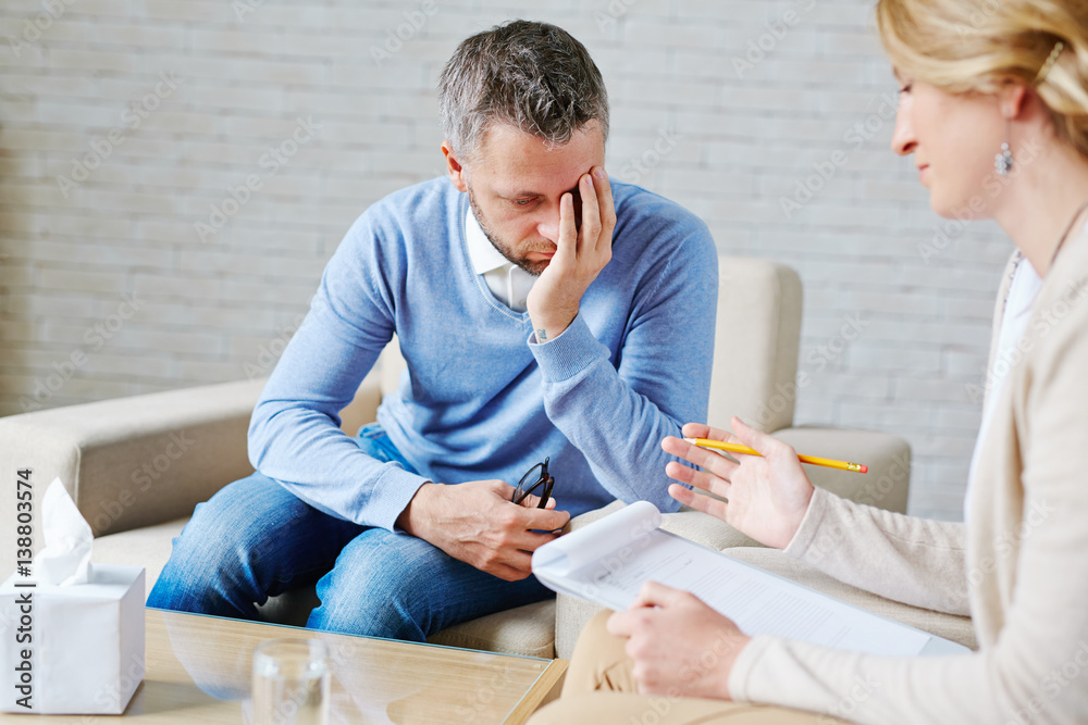 Ashamed bearded patient covering face with one hand while telling psychiatrist about his problems Ashamed bearded patient covering face with one hand while telling psychiatrist about his problems