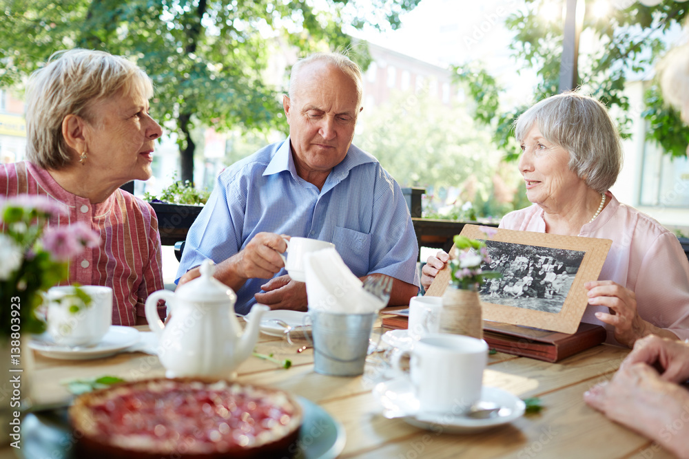 Senior friends gathered together in outdoor cafe and listening attentively to woman in pink blouse with old black-and-white photo in hands