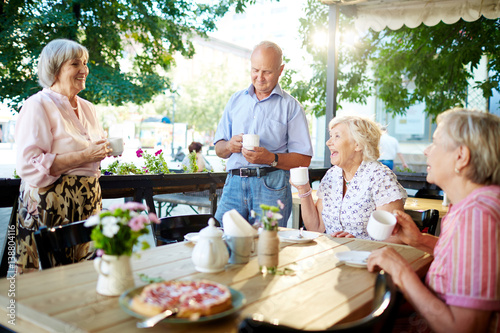 Cheerful senior friends celebrating holiday in outdoor cafe while one of them making toast, teapot and tasty pie standing on wooden table © pressmaster