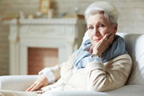 Pretty elderly woman in white shirt and beige cardigan leaning on elbow and looking at camera with slight smile