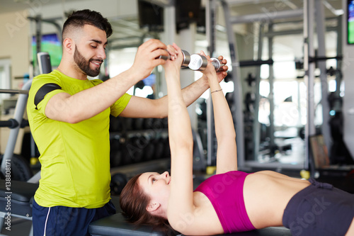 Handsome personal trainer helping young girl do correct dunbbell excercise on  flat bench photo