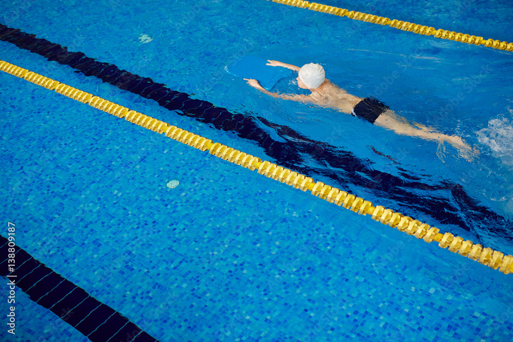 Top view image of unrecognizable school age boy swimming fast on pool lane separated by yellow lines in clear blue water