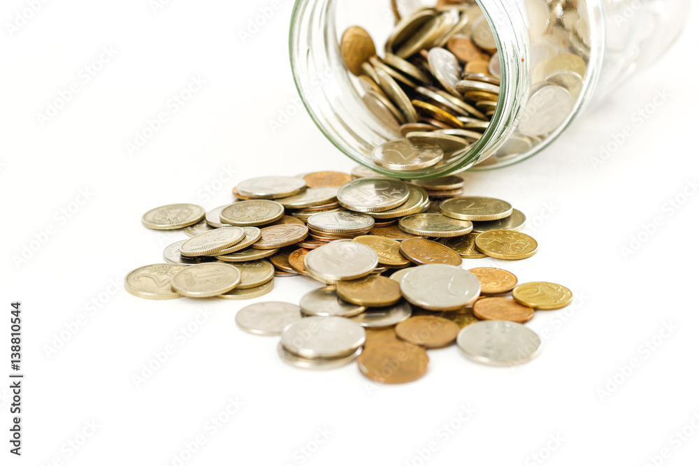 Close up coins in glass jar on white table. Coins scattered around. Isolated on white background. Close up coins in glass jar on white table. Coins scattered around. Isolated on white background.