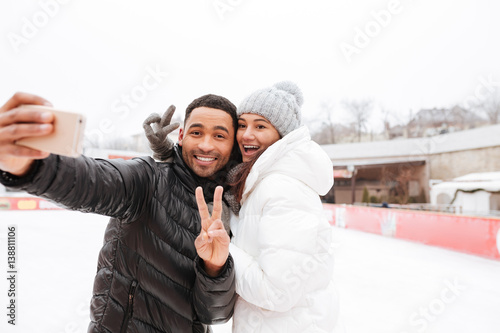 Smiling loving couple make selfie by phone and peace gesture.