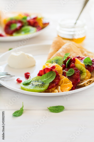 Thin crepes with fresh kiwi, orange, sicilian orange, honey, cream and mint leaves on white rustic wooden table. Selective focus