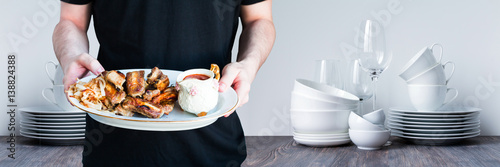 Young man with barbecued spareribs, pork ribs, tomatoe sauce and roast onion slices on white porcelain dish. Restaurant kitchen white background. Selective focus.
