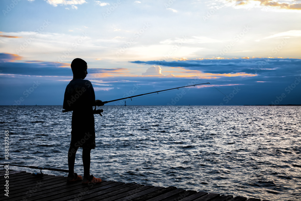 Silhouette of the boy fishing on wooden bridge extended into the sea on sunset.