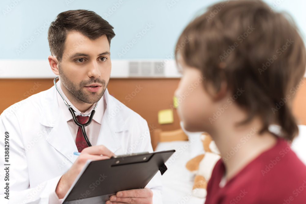 Man doctor with clipboard looking at little boy patient in hospital Man doctor with clipboard looking at little boy patient in hospital