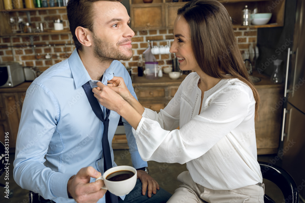 smiling businesswoman helping husband with shirt during breakfast