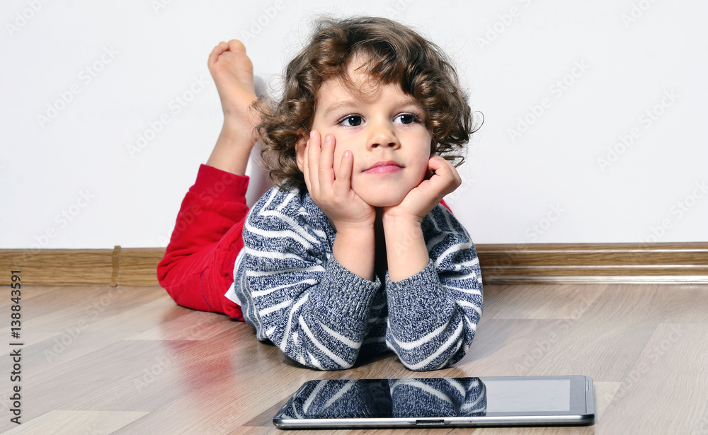 Beautiful kid playing games on a tablet. Boy sitting on the floor and looking on the display of a tablet watching cartoons. Beautiful kid playing games on a tablet. Boy sitting on the floor and looking on the display of a tablet watching cartoons.
