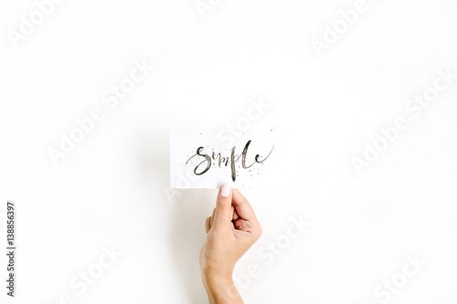 Minimal pale composition with girl's hand holding card with word Simple written in calligraphic style on paper on white background. Flat lay, top view