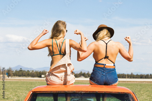 Two women sitting side by side on car roof, flexing arm muscles 