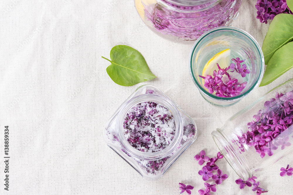 Glass jar of lilac flowers in sugar, glass and pitcher of lilac water with lemon and branch of fresh lilac on white linen tablecloth. Flat lay Glass jar of lilac flowers in sugar, glass and pitcher of lilac water with lemon and branch of fresh lilac on white linen tablecloth. Flat lay