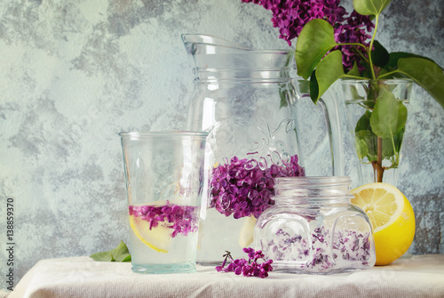 Glass jar of lilac flowers in sugar  glass and pitcher of lilac water with lemon  and branch of fresh lilac on white linen tablecloth with blue textured wall at background.