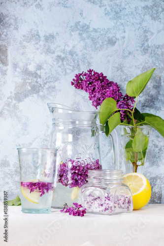 Glass jar of lilac flowers in sugar, glass and pitcher of lilac water with lemon, and branch of fresh lilac on white linen tablecloth with blue textured wall at background.