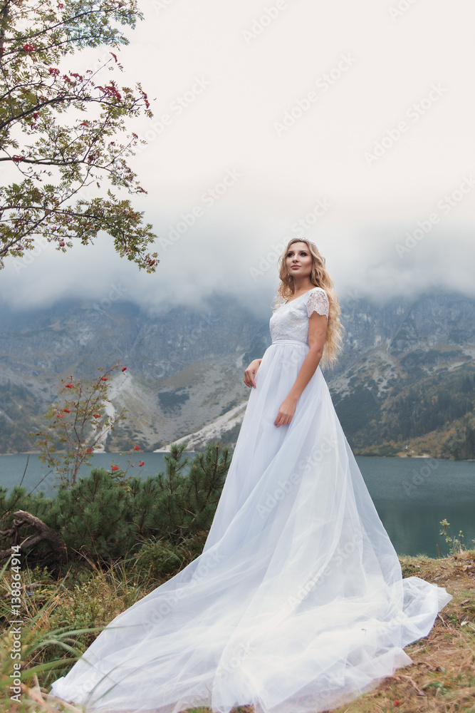 beautiful gentle girl bride in the air fairy blue wedding dress with luxurious curls in the mountains near the lake with a crown utterly