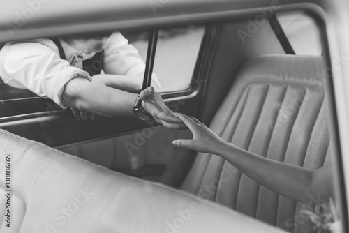 Groom reaches out bride sitting in an old car, clock hand, black-and-white photo