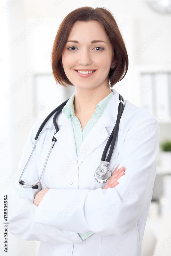 Young brunette female doctor standing with arms crossed and smiling at hospital. Health care, insurance and help concept. Physician ready to examine patient