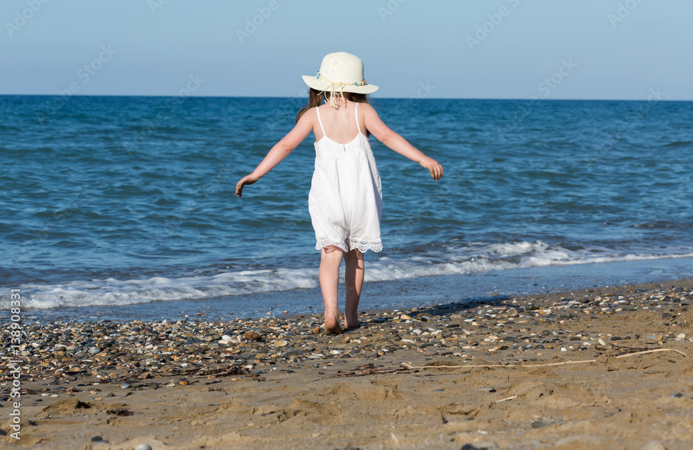 Little girl in white dress walking alone the sea, playing on the seashore at the sunset