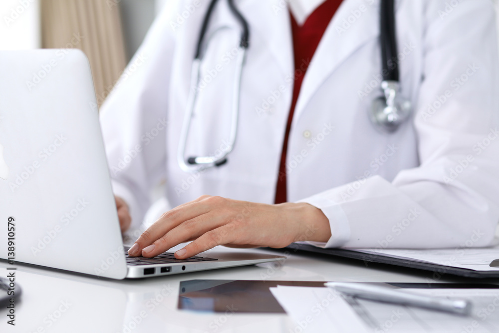 Close up of unknown female doctor typing on laptop computer while sitting  at the table. Medical staff, compulsory work concept