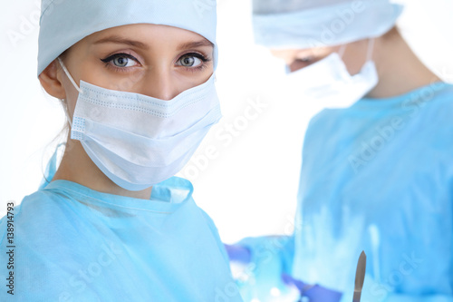 Close up of surgeon woman looking at camera while colleagues performing in background in operation room, isolated. Surgery and medicine concept
